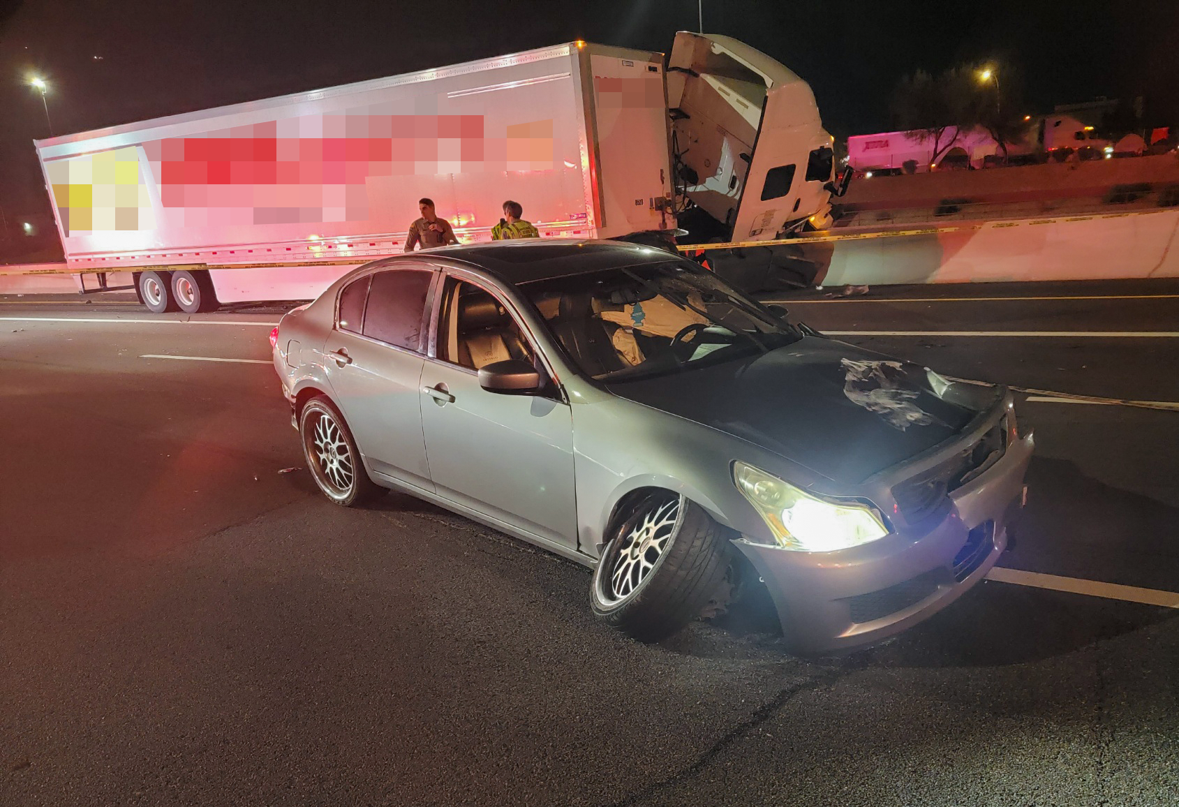 A gray sedan with heavy front end damage sits in the middle of highway lanes beside a white tractor-trailer that has crashed into a median wall