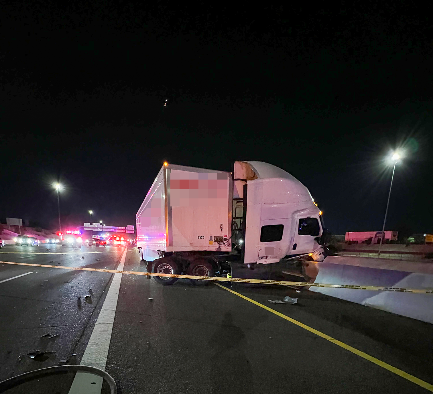 A crashed white tractor-trailer partially into the highway median barrier wall with the cab turned sideways