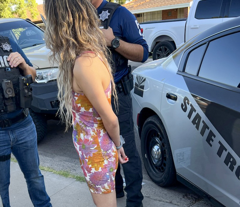A woman in handcuffs stands next to a police vehicle with uniformed police detectives next to her
