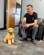 An AZDPS detective sits in an office chair next to a small tan dog on a leash