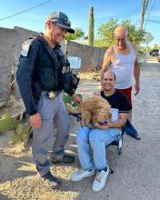  A small tan dog sits in the lap of man in a folding chair while a SWAT trooper and another man look on and smile