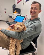 A photograph of a small, tan dog sitting in the lap of an AZDPS detective in an office