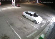Screenshot from a gas station camera showing a white car parked in front of a store. A man is visible in the driver's seat.