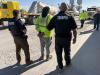 Two troopers stand behind a man wearing a high-visibility yellow shirt with his hands in handcuffs